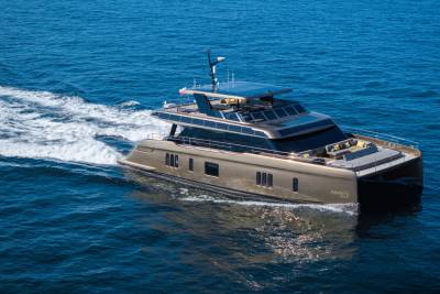 A sleek, golden catamaran yacht glides through calm blue water, leaving a white wake behind it, with a clear sky visible in the distance.