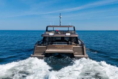 A modern catamaran yacht named "ASANTE" cruises through calm blue waters, leaving a white wake behind it under a clear sky.