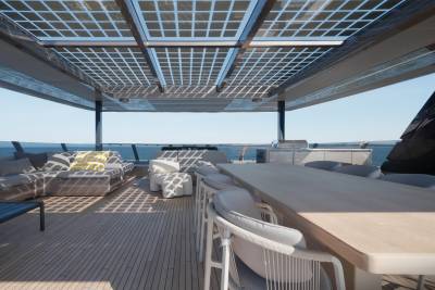 A spacious yacht deck with a dining table and lounge area in the foreground is set against a backdrop of the ocean and a clear sky.