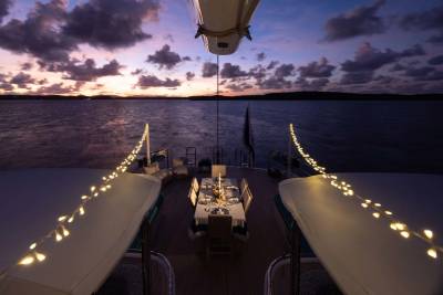 A beautifully set dining table on a yacht's deck is illuminated by fairy lights, with a dramatic sunset over the ocean in the background.