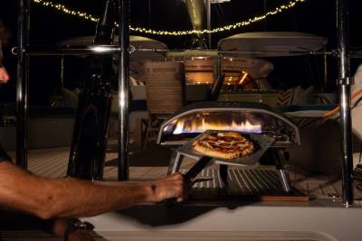 A person uses a pizza peel to place a pizza into a portable oven on a boat deck, with string lights illuminating the background.