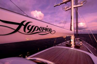 A large white sail with the name "Hyperion" in black script dominates the foreground, while the background features a tranquil ocean under a vibrant purple and pink sunset sky.