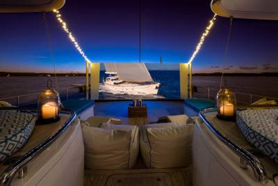 Lanterns and cushions adorn the deck of a yacht in the foreground, while a large screen displays a sailboat on the water against a twilight sky in the background.