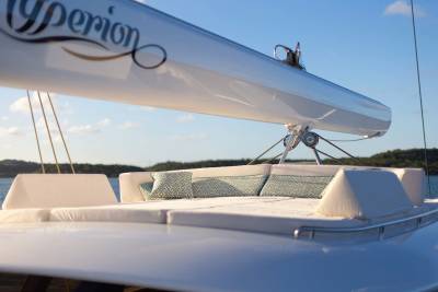 A white sailboat mast with the name "Hyperion" curves over a plush seating area on a yacht's deck, with a calm blue sea and distant green hills under a clear sky in the background.