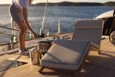 A person serves a drink and food on a yacht's deck with a lounge chair in the foreground and a calm sea in the background.