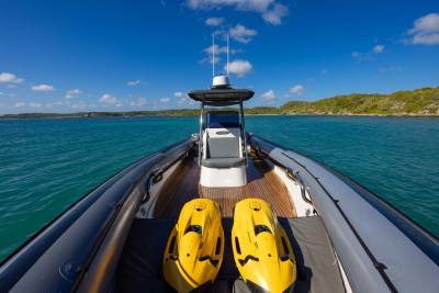 Two yellow sea scooters rest on the deck of a rigid inflatable boat, with a calm turquoise sea and a lush green coastline under a bright blue sky in the background.