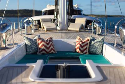 Two patterned pillows rest on teal cushions on the deck of a yacht, with lounge chairs and a larger vessel visible in the background under a clear sky.