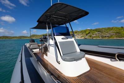A modern inflatable boat with a wooden deck sits on calm turquoise water, with a lush green, hilly coastline under a bright blue sky in the background.