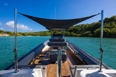 A sleek, modern boat with a dark canopy sits on calm, turquoise water, with lush green hills and a bright blue sky in the background.