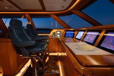 Luxurious leather captain's chairs and a navigation chart sit in the foreground of a yacht's bridge, while a dark, twilight sky is visible through the large windows in the background.