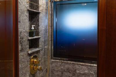 A modern shower with marble walls and gold fixtures is visible in the foreground, while a large, frosted window with a blue light emanates from behind it in the background.