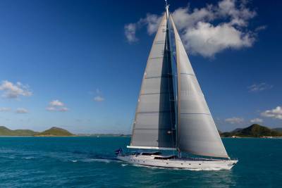 A large white sailboat with full sails glides across turquoise water under a bright blue sky with scattered clouds, with green islands visible in the distance.