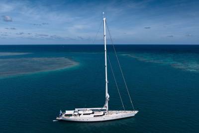 A large white sailboat with tall masts glides through clear turquoise water, with coral reefs visible beneath the surface and a vast blue sky above.