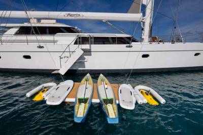 Four paddleboards and two yellow jet skis are docked on a wooden platform in the foreground, with a large white yacht docked behind them.