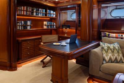 A luxurious wood-paneled office with a desk and laptop in the foreground is set against a background of bookshelves and porthole windows.