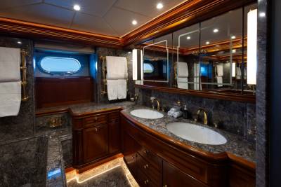 A luxurious yacht bathroom features a double vanity with marble countertops and wooden cabinetry in the foreground, while the background shows a large mirror reflecting the space and an oval window with a view of the sky.