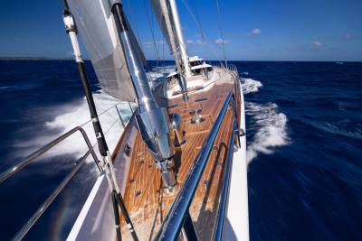 A sleek sailboat with a wooden deck cuts through the deep blue ocean, leaving a white wake under a clear sky.