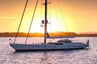 A large white sailboat rests on calm water in the foreground, silhouetted against a vibrant orange sunset over distant hills in the background.