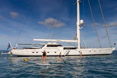 Four people paddleboard in the foreground with a large white yacht anchored in the background under a blue sky with scattered clouds.