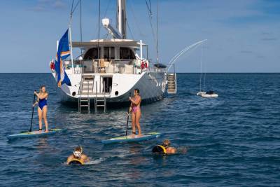 Two women paddleboard and two people snorkel in the foreground, with a large white yacht anchored in the blue ocean behind them.
