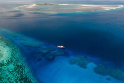 A luxurious yacht floats in the deep blue ocean, surrounded by vibrant coral reefs in the foreground and a distant, sandy island in the background.