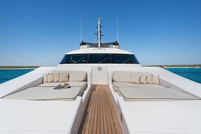 Two lounge areas with beige cushions and pillows sit on the white deck of a yacht, with a clear blue sky and ocean in the background.