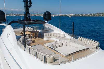 A luxury yacht deck with white lounge seating and a wooden dining area sits against a backdrop of deep blue ocean and a distant coastal city.