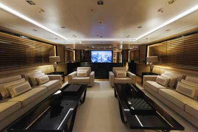 Two long white sofas and black coffee tables face each other in the foreground of a luxurious lounge, with a large screen displaying a city skyline in the background.