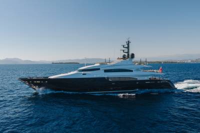 A sleek, dark-hulled luxury yacht cruises through deep blue waters against a backdrop of distant mountains and a clear sky.