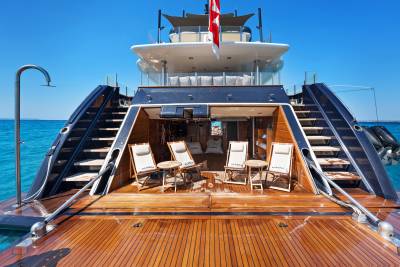 A luxury yacht's wooden deck with lounge chairs sits in the foreground, set against a clear blue sky and ocean background.
