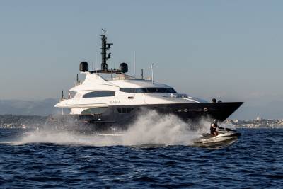 A small motorboat kicks up white spray in the foreground, while a large luxury yacht named "ALASKA" cruises across the blue water against a backdrop of distant mountains and a city skyline.