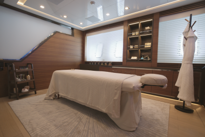 A white massage table sits on a light-colored rug in a modern spa room with wood-paneled walls, shelving, and blinds.