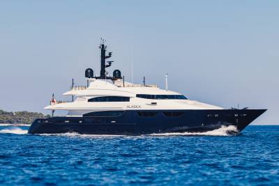A large, dark-hulled luxury yacht named "ALASKA" cruises through deep blue water under a clear, pale sky.