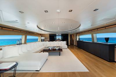 A spacious white sectional sofa and coffee table sit in the foreground of a luxury yacht interior, with panoramic ocean views visible through large windows in the background.
