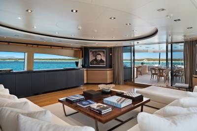 A modern yacht lounge with white sofas and a coffee table in the foreground opens up to a dining area and a scenic ocean view in the background.