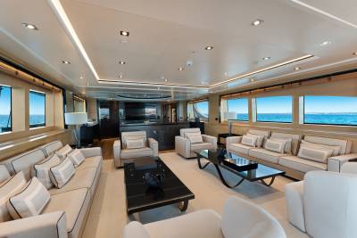 A modern yacht lounge features plush white seating and black coffee tables in the foreground, with a sleek bar area and panoramic ocean views through large windows in the background.