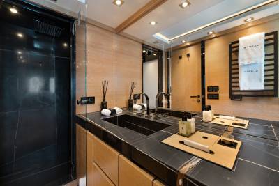 A black marble vanity with toiletries and a sink sits in the foreground, while a dark shower stall and tan walls are visible in the background.