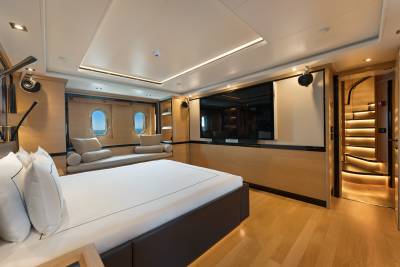 A large bed with white linens occupies the foreground of a modern yacht cabin, while the background features a built-in sofa by two portholes, a wall-mounted television, and a lit staircase.