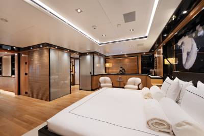 A large bed with white linens occupies the foreground of a modern, wood-paneled bedroom featuring a seating area and sleek architectural lighting in the background.