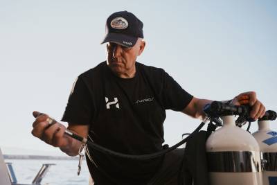 A man in a black t-shirt and baseball cap prepares diving equipment in the foreground against a clear blue sky and ocean background.