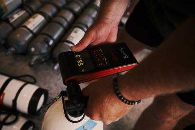 A person's hands use a digital analyzer to check the gas levels of a white tank in the foreground, with several grey diving cylinders lined up in the background.