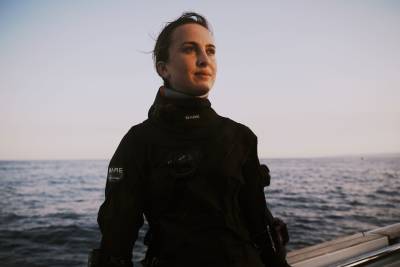A woman wearing a black drysuit stands in the foreground against a background of a calm ocean and a clear sky.