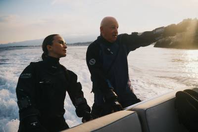 A man and woman in wetsuits stand on a boat, with the man pointing toward a distant coastline across the churning ocean water.