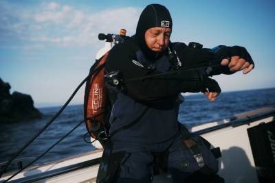 A scuba diver in a black hood and blue wetsuit prepares his gear on a boat against a backdrop of blue ocean and sky.