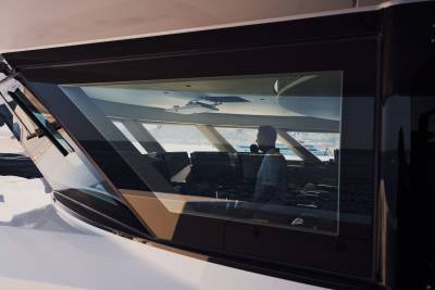A man is seen through the glass window of a white yacht, with a body of water and distant hills in the background.
