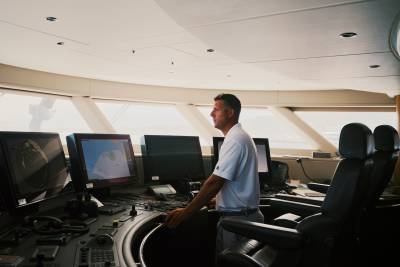 A man in a white shirt stands at a high-tech navigation console in the foreground, with the bright horizon visible through the bridge windows in the background.