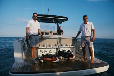 Two men in white shirts and grey shorts stand on the wooden deck of a boat equipped with diving gear, set against a calm blue ocean and clear sky.