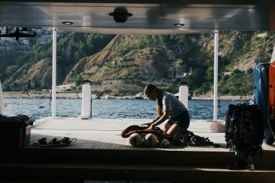 A woman kneels on the wooden deck of a boat preparing gear, with a scenic coastline and blue water in the background.