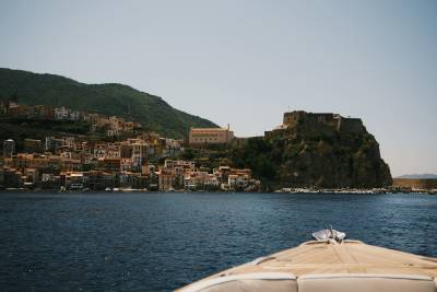 The bow of a boat sits in the foreground of a deep blue sea, with a hillside coastal town and a fortress overlooking the water in the background.
