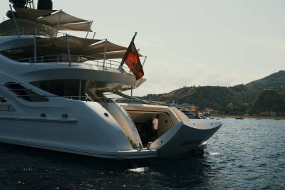 A large white luxury yacht with a red flag floats in the foreground against a backdrop of calm blue water and distant green mountains.
